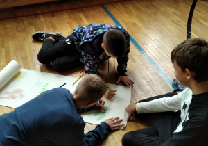 Na podłodze rozłożony jest duży biały karton. Dwóch chłopców rysuje elementy swojego plakatu a trzeci trzyma papier i obserwuje prace swoich kolegów.