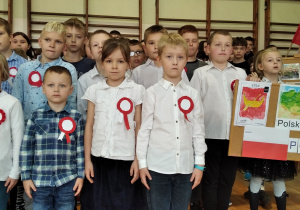 Na zdjęciu dzieci są ubrane w odświętne stroje tuż przed odśpiewaniem hymnu. Mają na sobie kotyliony. Dwóch uczniów z klasy pierwszej trzyma tablicę, na której są ilustracje mapy Polski oraz biało- czerwona flaga naszego kraju, a także herb Łodzi (patrząc z prawej strony).