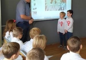 Uczeń z klasy ósmej zadaje pytania z ,,Katechizmu polskiego dziecka" dziewczynkom z klasy pierwszej. Dzieci ubrane są w odświętne stroje. Mają na sobie przyczepione kotyliony.