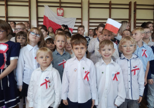Grupa dzieci w strojach galowych z biało-czerwonymi kotylionami podczas szkolnej uroczystości patriotycznej. W tle widoczna flaga Polski.